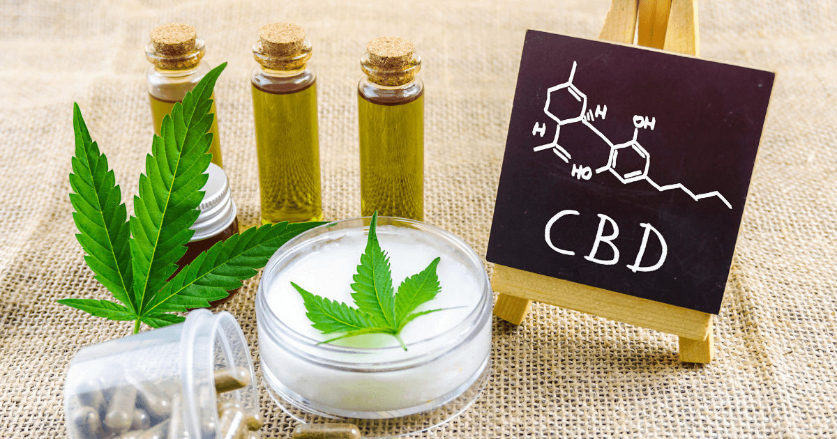 Cancer and CBD: Three vials of CBD oil next to a cannabis leaf, a topical with a cannabis leaf against the white cream. A small blackboard sits next to this with CBD and the chemical equation on it