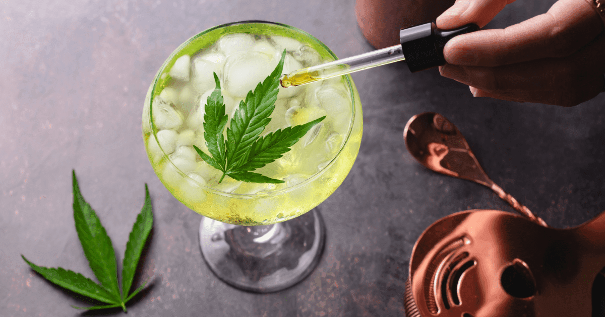 CBD and alcohol: A cocktail glass with a yellow liquid sits next to cocktail making eupiment. A hand reaches over and squeezes CBD oil from a dropper into the liquid which is topped by a cannabis leaf