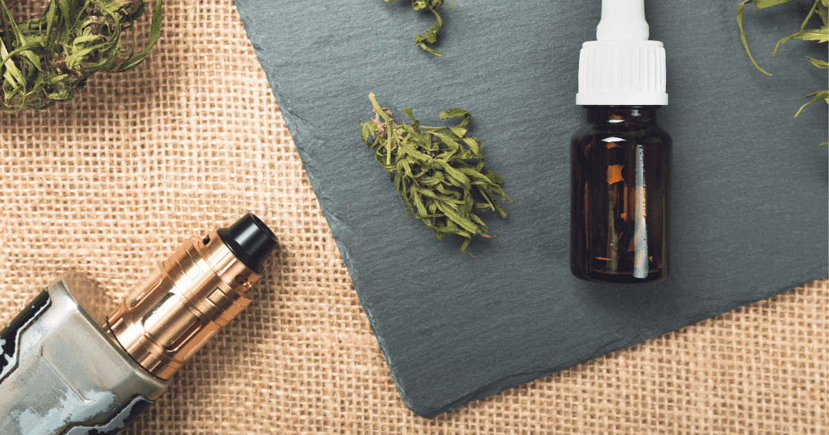 Vaping study: A bottle of oil lies on its side on a grey background. This is resting on a sandy coloured background. There is a vape lying to the left of the image and assorted cannabis buds dotted throughout