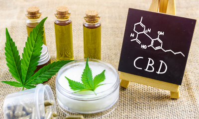 Cancer and CBD: Three vials of CBD oil next to a cannabis leaf, a topical with a cannabis leaf against the white cream. A small blackboard sits next to this with CBD and the chemical equation on it
