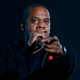 Jay-Z: A photo of the rapper Jay-Z against a black background. He is holding a microphone and rapping into it