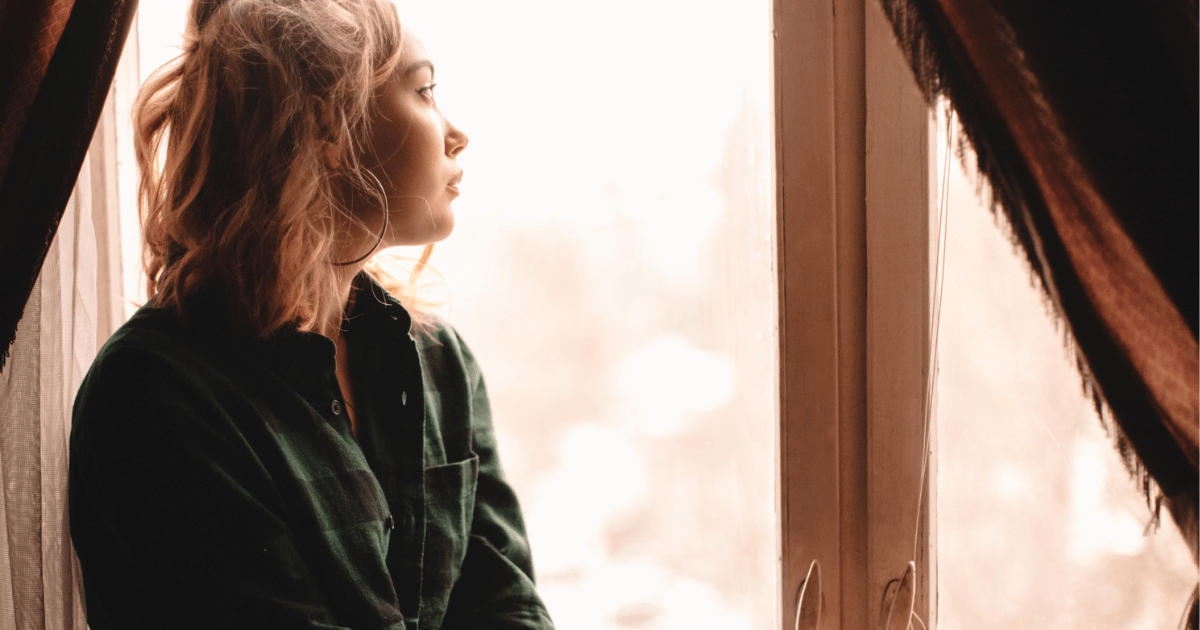 Motivation: A woman with blonde hair and a green jumper sits at a window looking outside with a bored expression