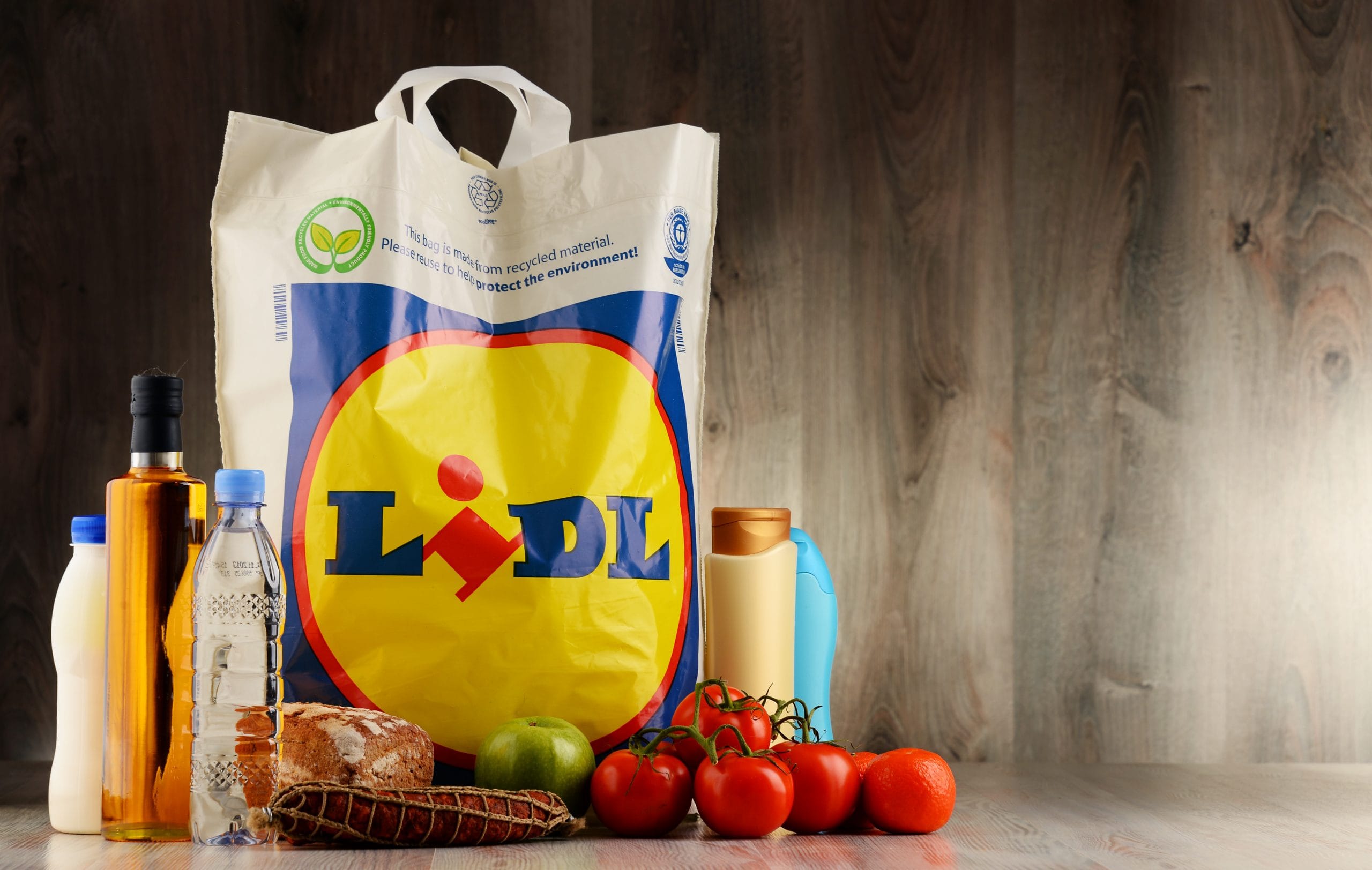 Lidl: A plastic shopping bag on a wooden surface surrounded by milk and oil bottles, tomatos, apples and food items