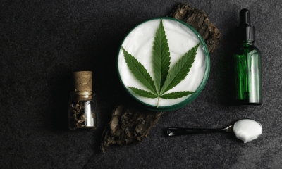 Skin condition: A white cream with a green cannabis leaf on top. It is surrounded by dark oil bottles and a spoon that has cream on it. This is on a dark background