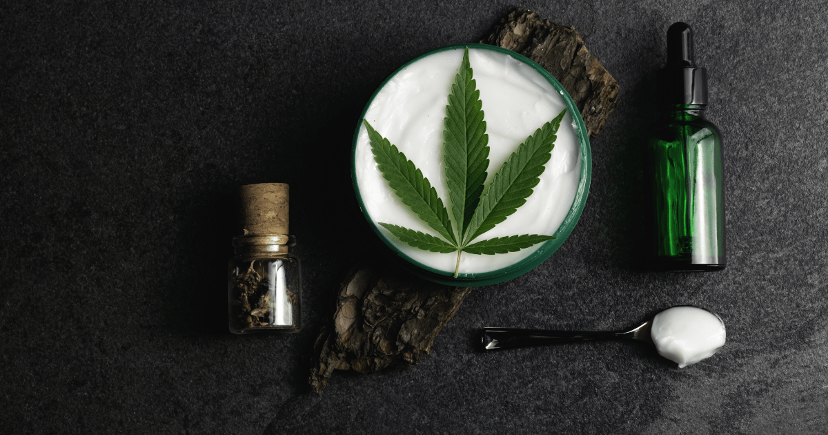 Skin condition: A white cream with a green cannabis leaf on top. It is surrounded by dark oil bottles and a spoon that has cream on it. This is on a dark background