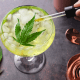 CBD and alcohol: A cocktail glass with a yellow liquid sits next to cocktail making eupiment. A hand reaches over and squeezes CBD oil from a dropper into the liquid which is topped by a cannabis leaf