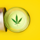 Seizures: A small jar of CBD topical gel on a yellow background. The lid off the jar and it has a cannabis leaf on the top of white cream